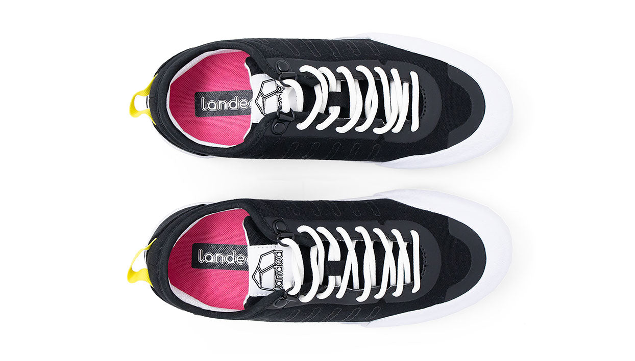 Landed The Overlook 1.0 black technical sneakers with white laces, pink insoles, and yellow heel pull tabs, top view.